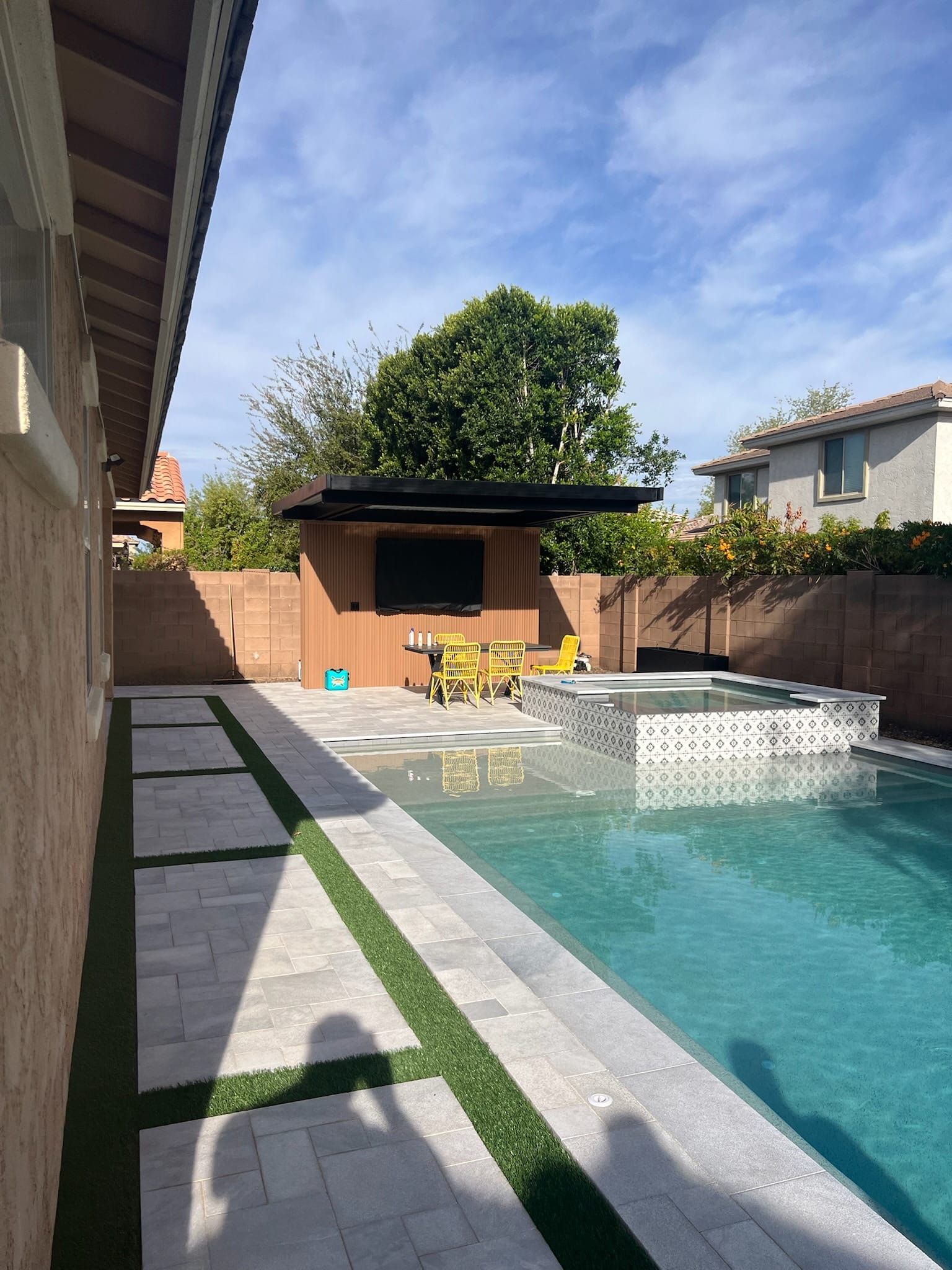 A pool and patterned-tile spa with a slatted-wood cabana, black pergola roof and yellow patio chairs on gray pavers.