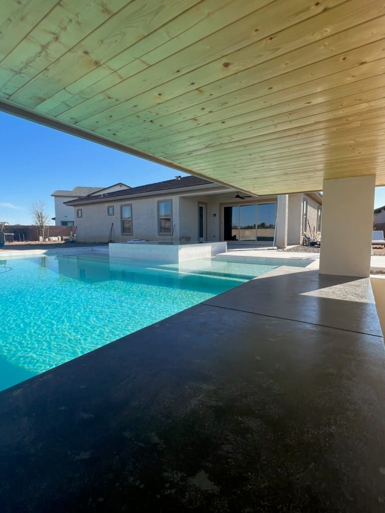 A pool and raised white-tile spa seen from under a wood-plank patio ceiling, with dark stained concrete decking in the foreground.