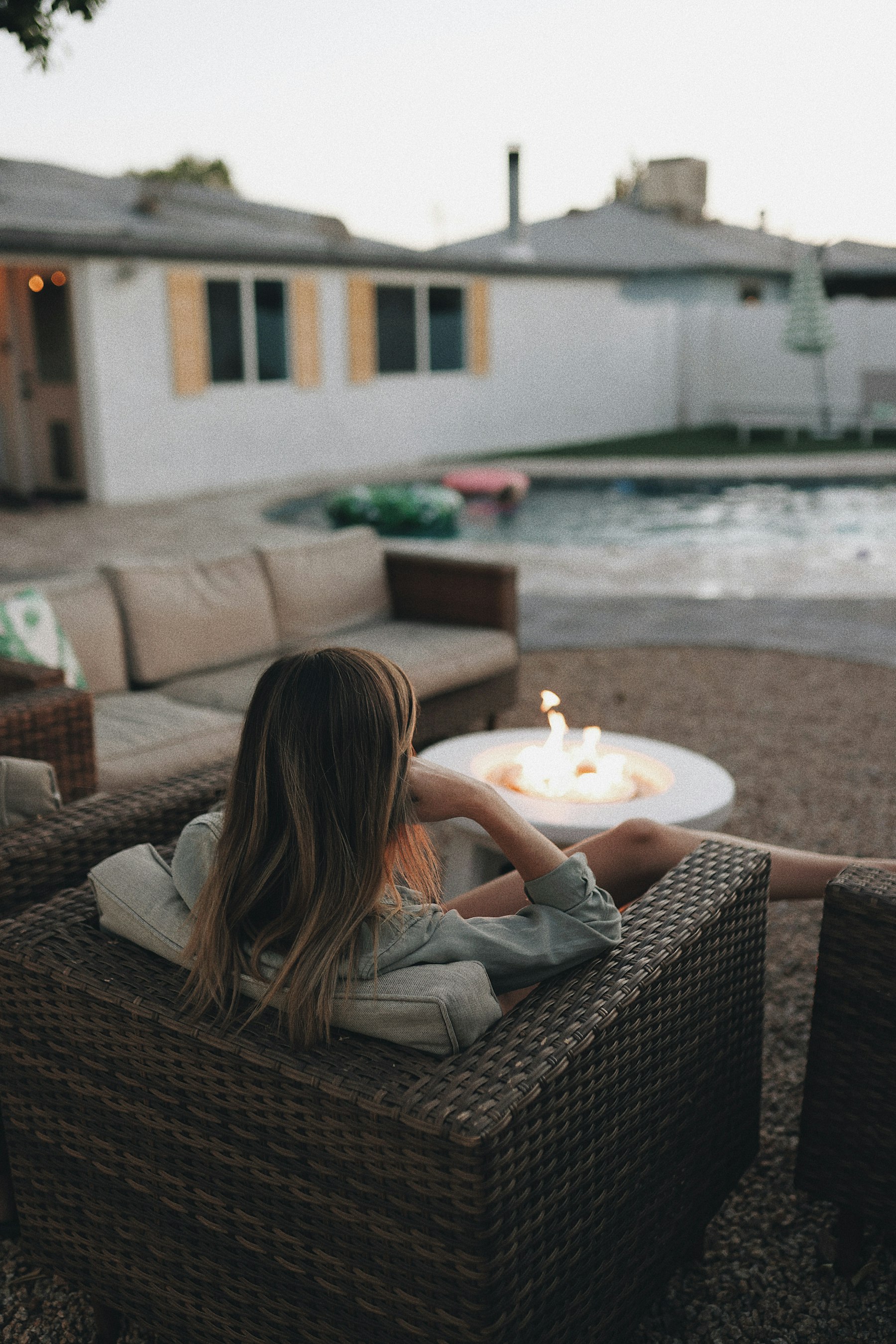 A round gas fire feature glowing beside a backyard pool at dusk, with block-wall home and gravel patio.