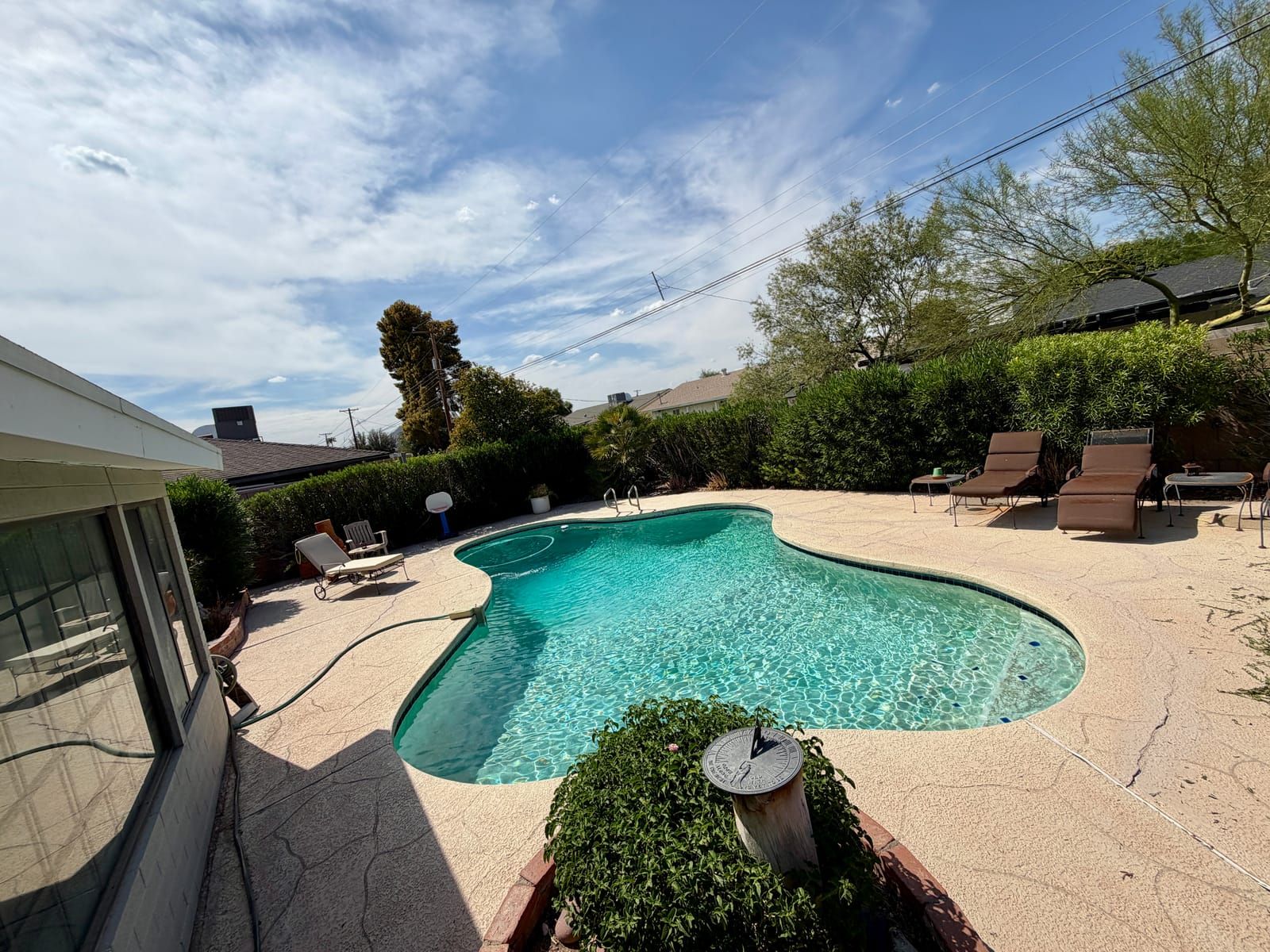 A freeform pool with flagstone-textured cool decking, framed by a tall oleander hedge, palo verde trees and desert mountains, with loungers and a sundial planter.