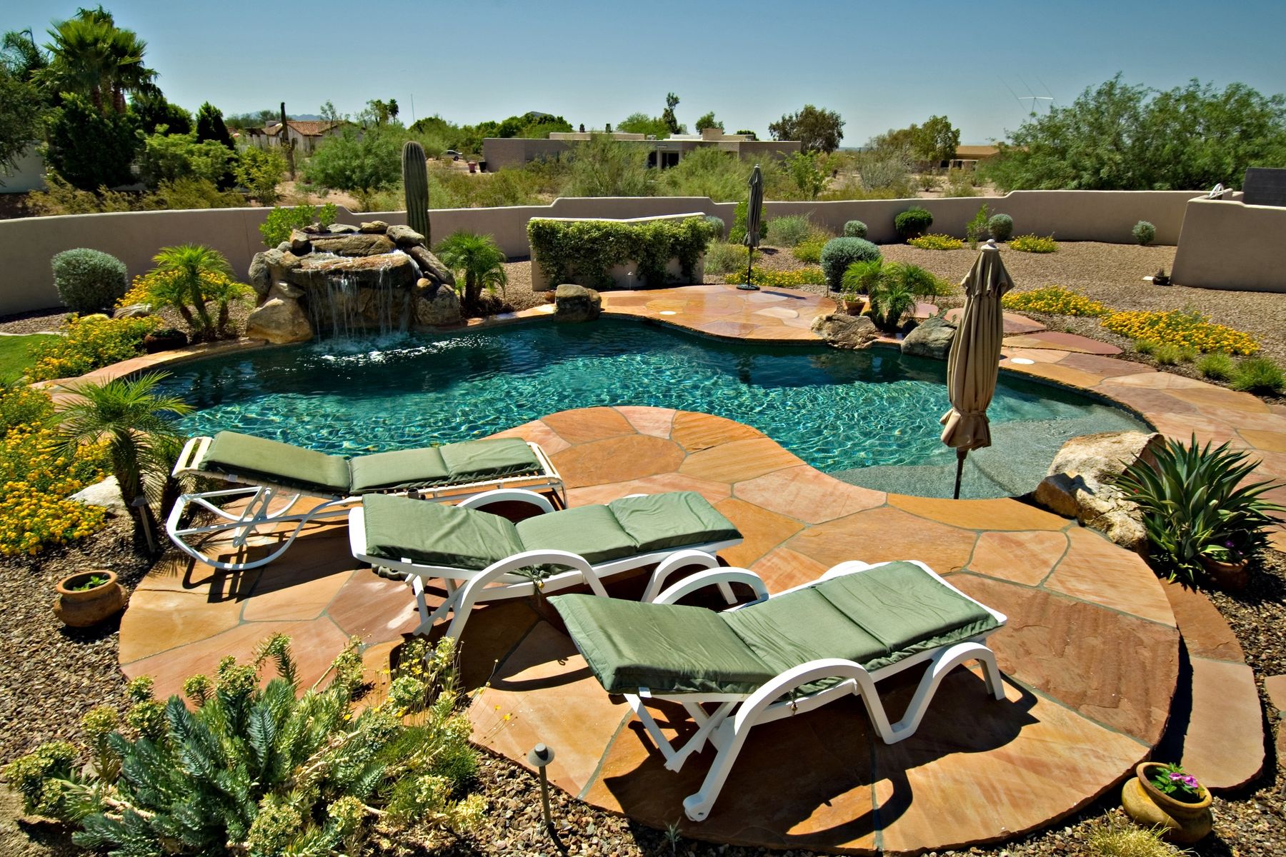 A freeform desert backyard pool with flagstone decking and a rock waterfall, framed by saguaro cactus, agave and xeriscape gravel, with a block perimeter wall and open Sonoran desert beyond.