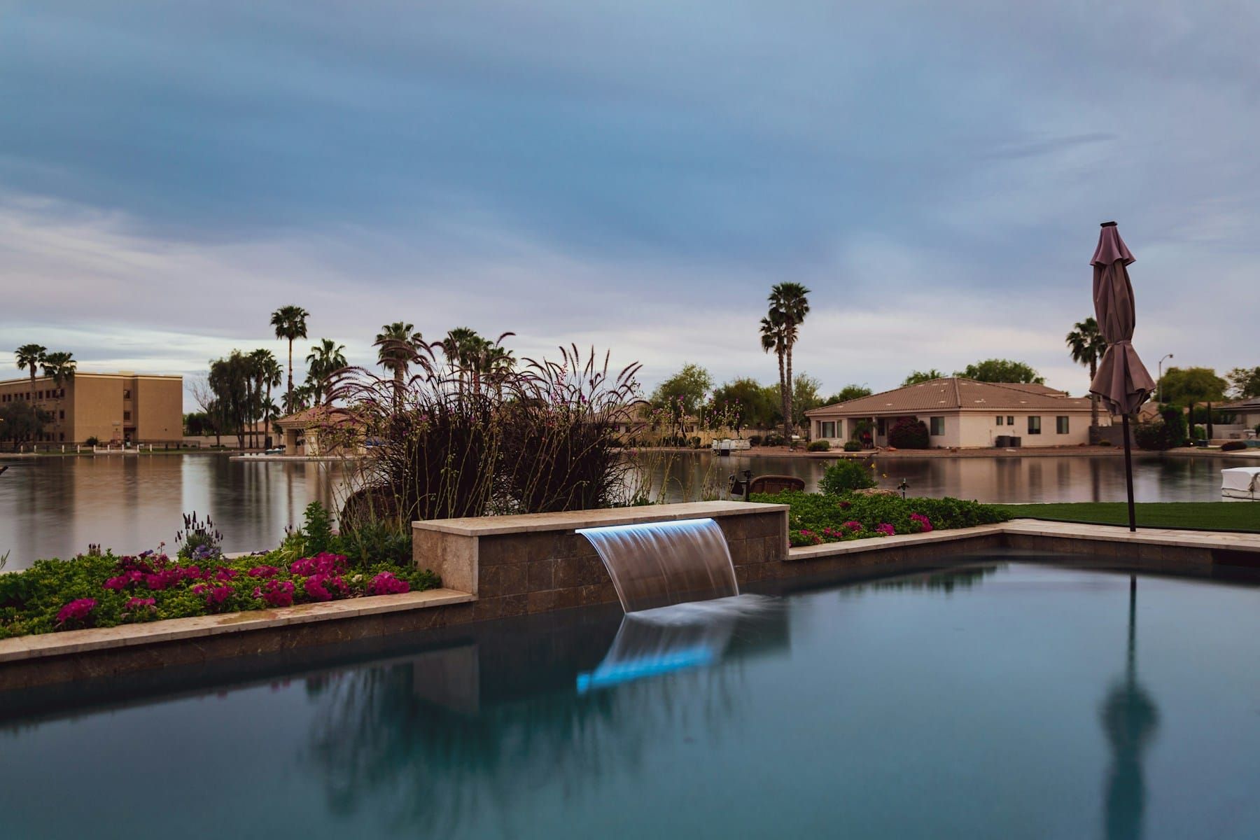 A lit sheet-descent water feature pouring from a travertine raised wall into a backyard pool at dusk, with a community lake, palms and stucco homes behind.