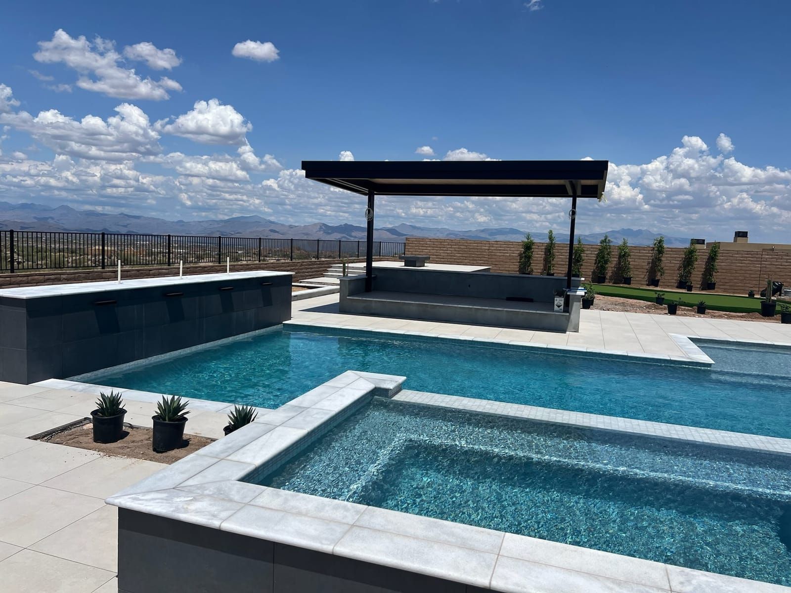 A modern pool and raised spa with marble coping and a covered bar pavilion, overlooking desert mountains under a blue sky.