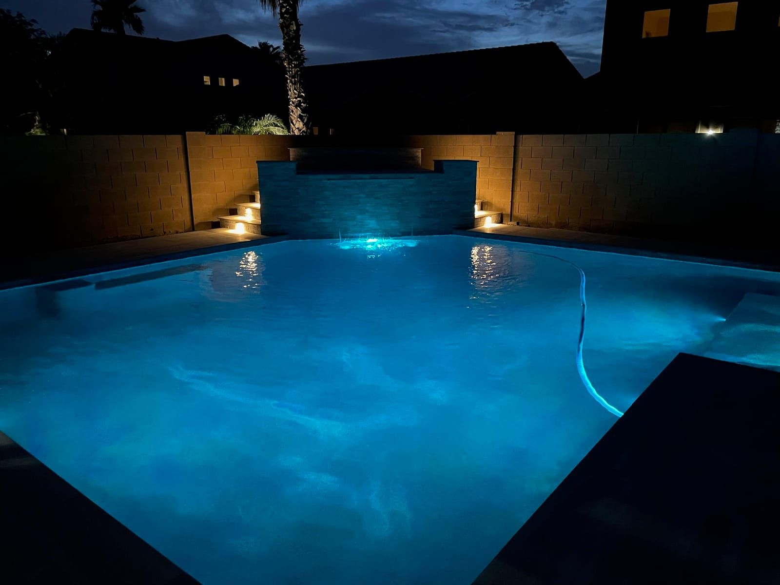 A pool glowing teal at night below a stacked-stone raised wall with a sheet waterfall, flanked by warm step lights on block walls.