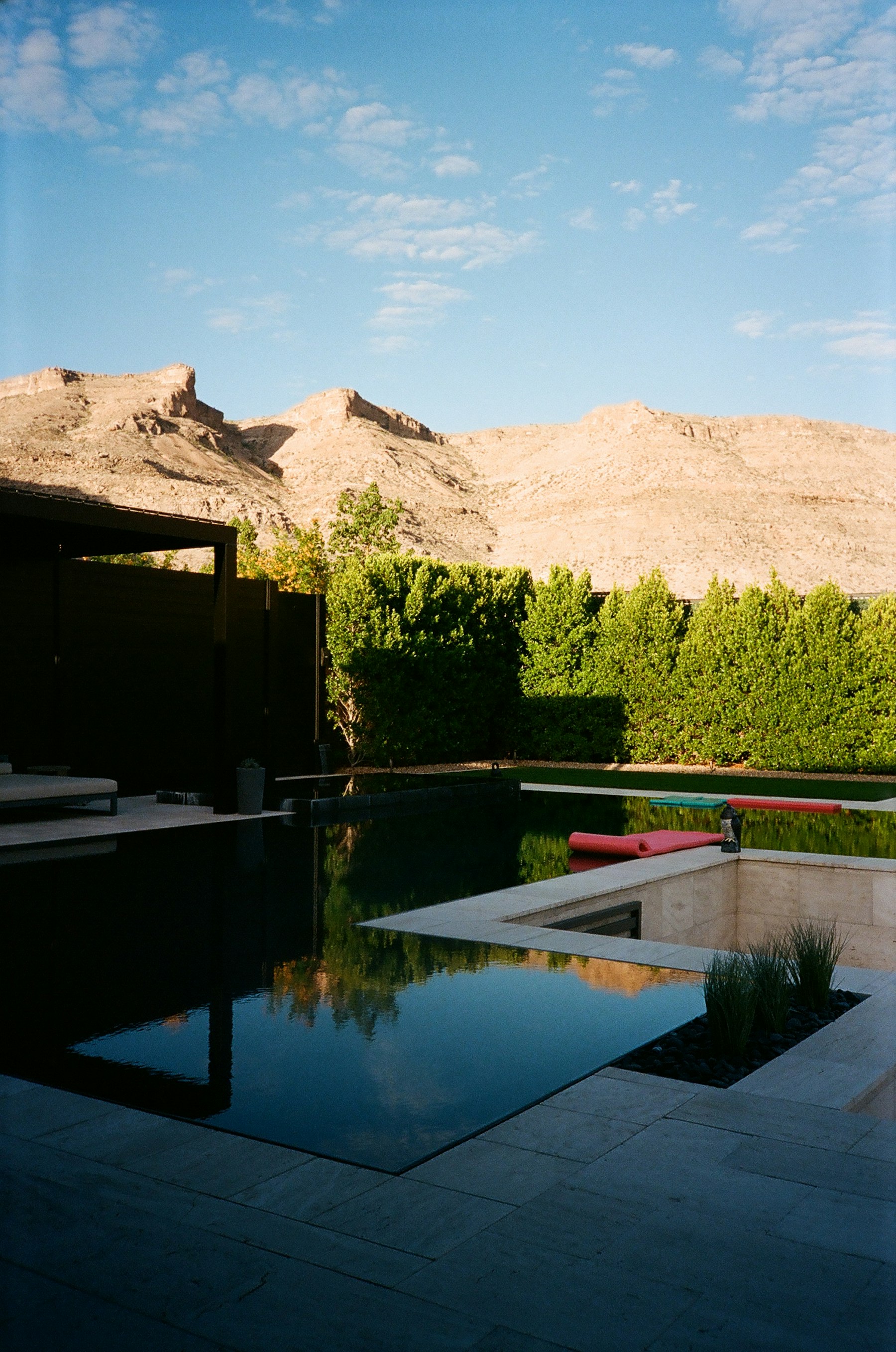 A dark-bottom pool with a raised spa and spillover, travertine decking and desert mountains behind a privacy hedge.