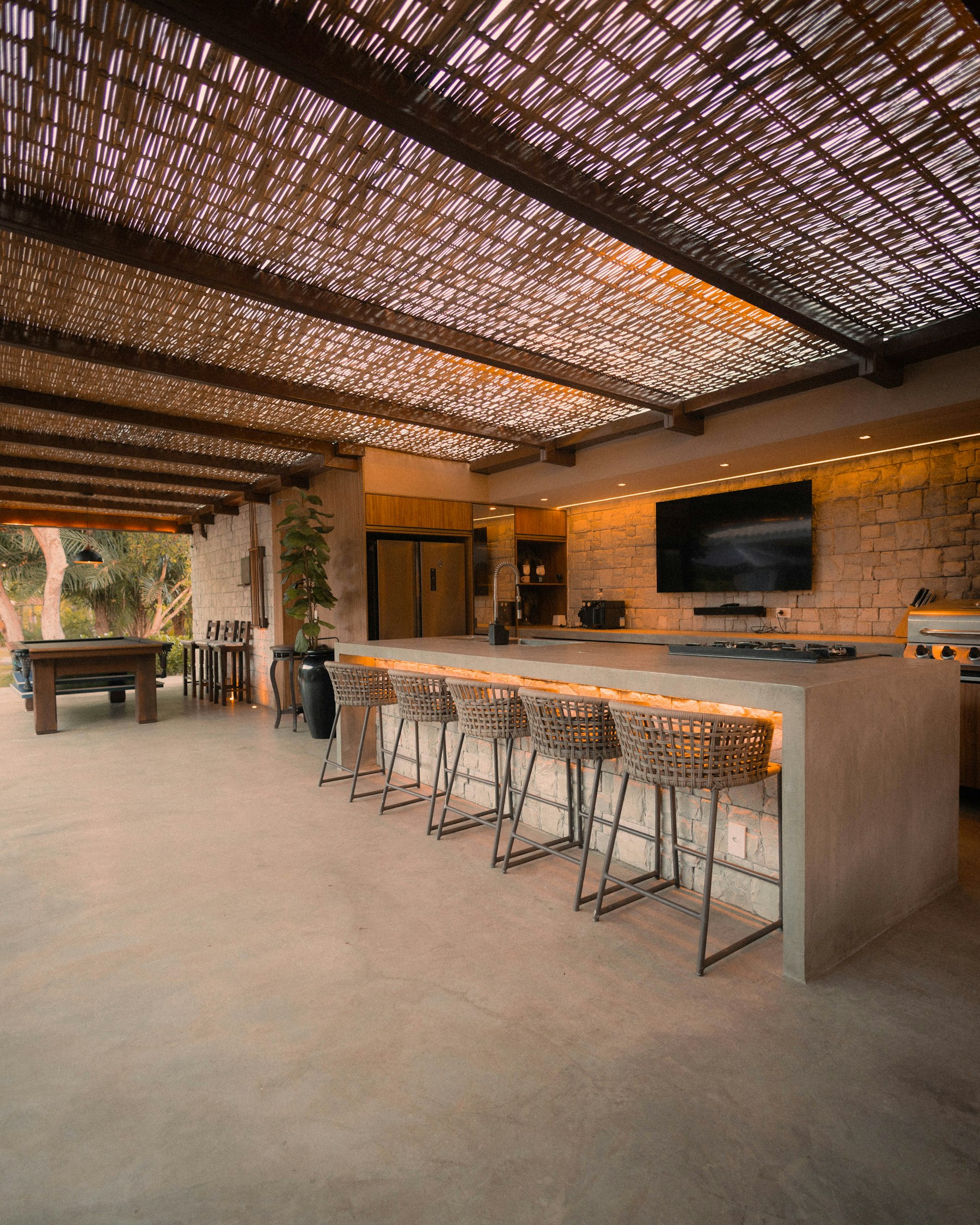 A ramada-shaded outdoor bar and kitchen with stone counter, bar stools and a built-in grill lit warmly at dusk.