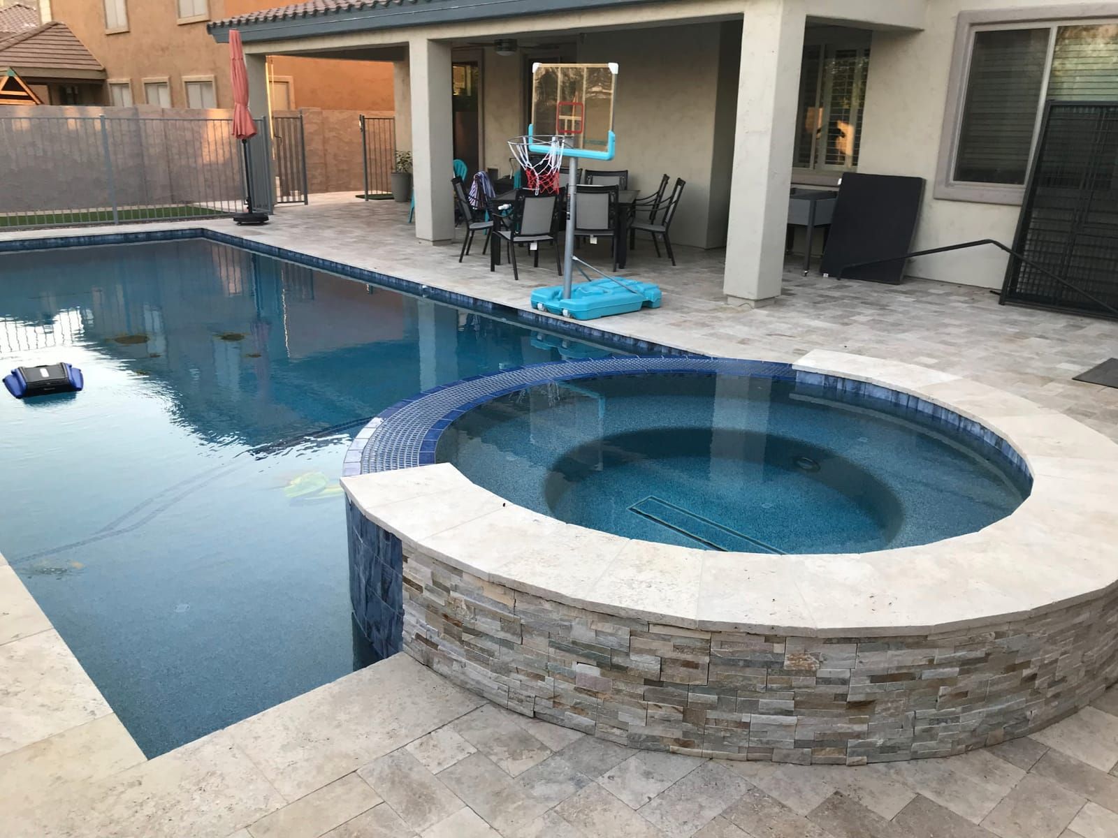 A round raised spa faced in stacked stone with blue glass waterline tile, spilling into a pool wrapped in travertine decking.