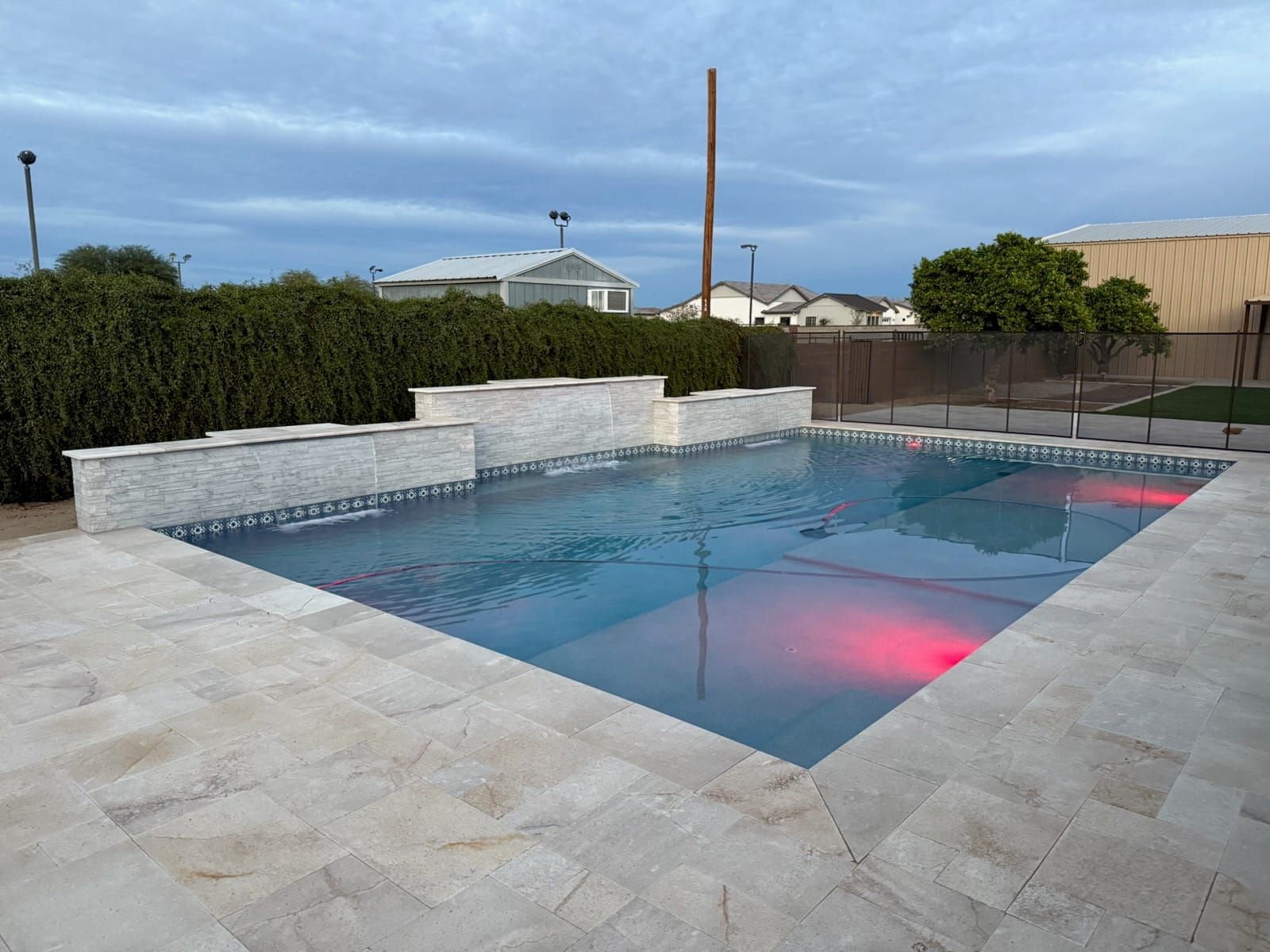 A rectangular pool at dusk with red underwater lights and raised stacked-stone waterfall walls behind a travertine deck.