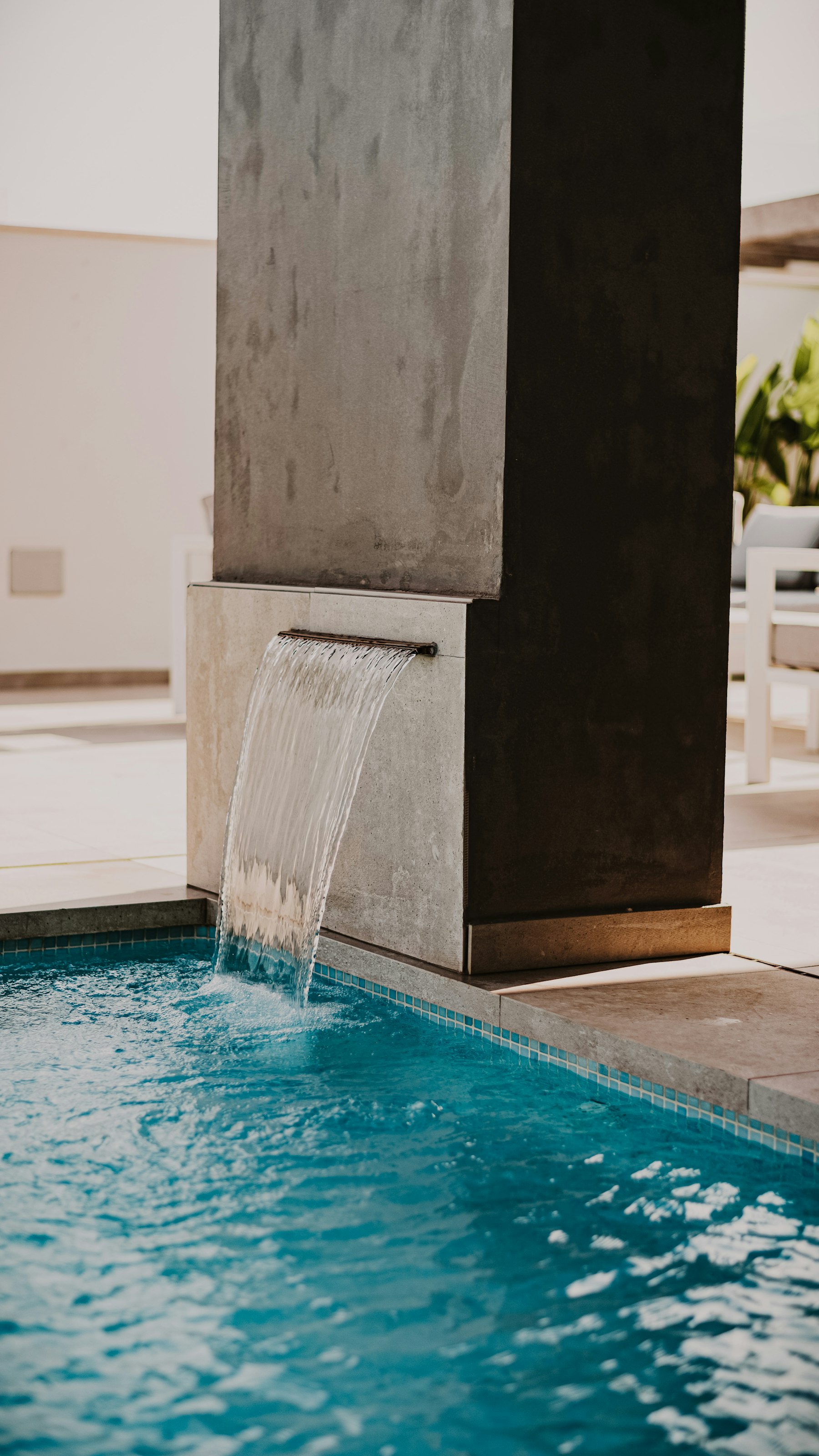 Close view of a sheet-descent water feature pouring from a concrete-and-stone wall into a tiled pool edge.