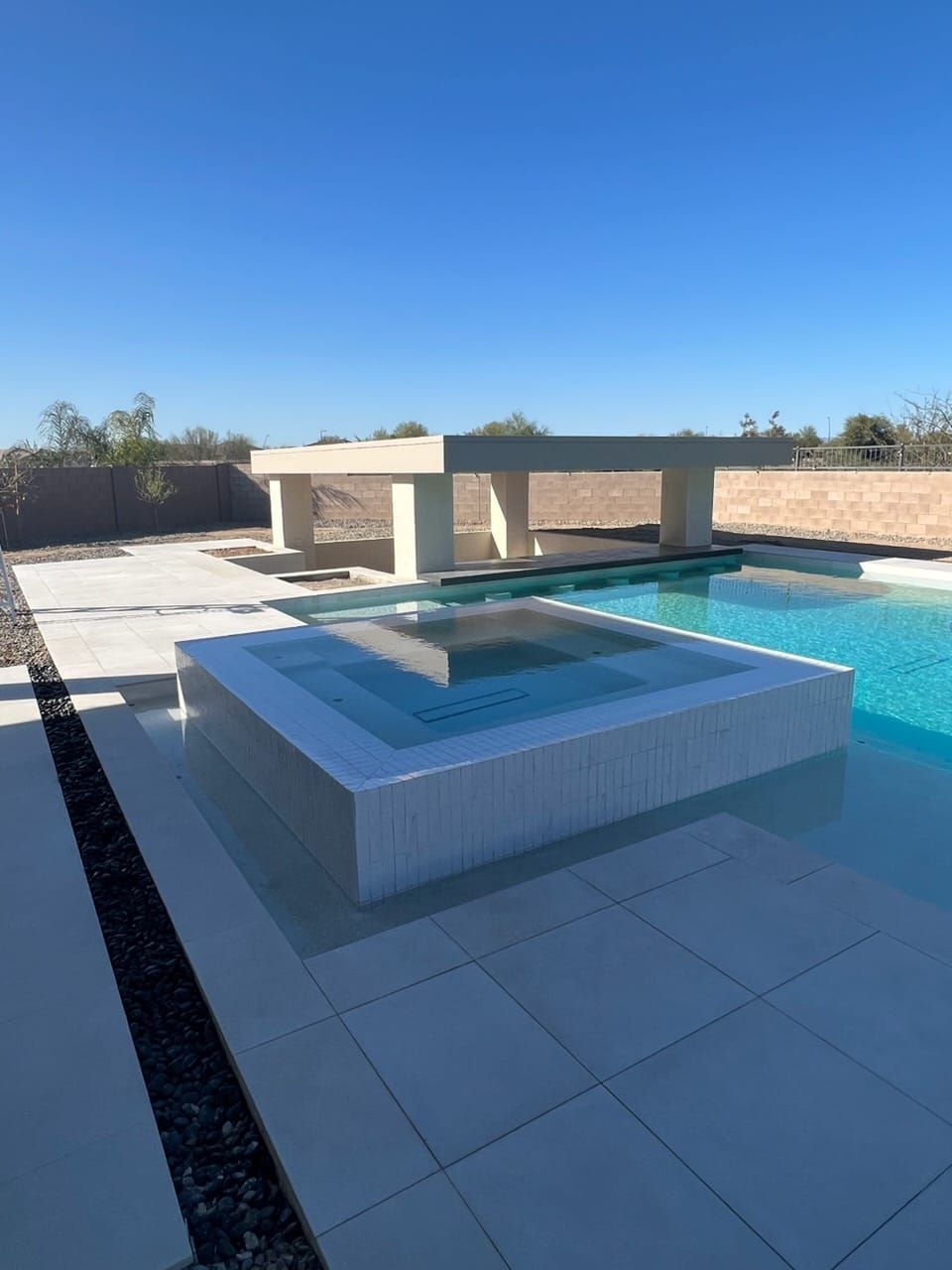 A square raised spa fully clad in white tile at the edge of a bright blue pool, with a shade pavilion and block wall behind.
