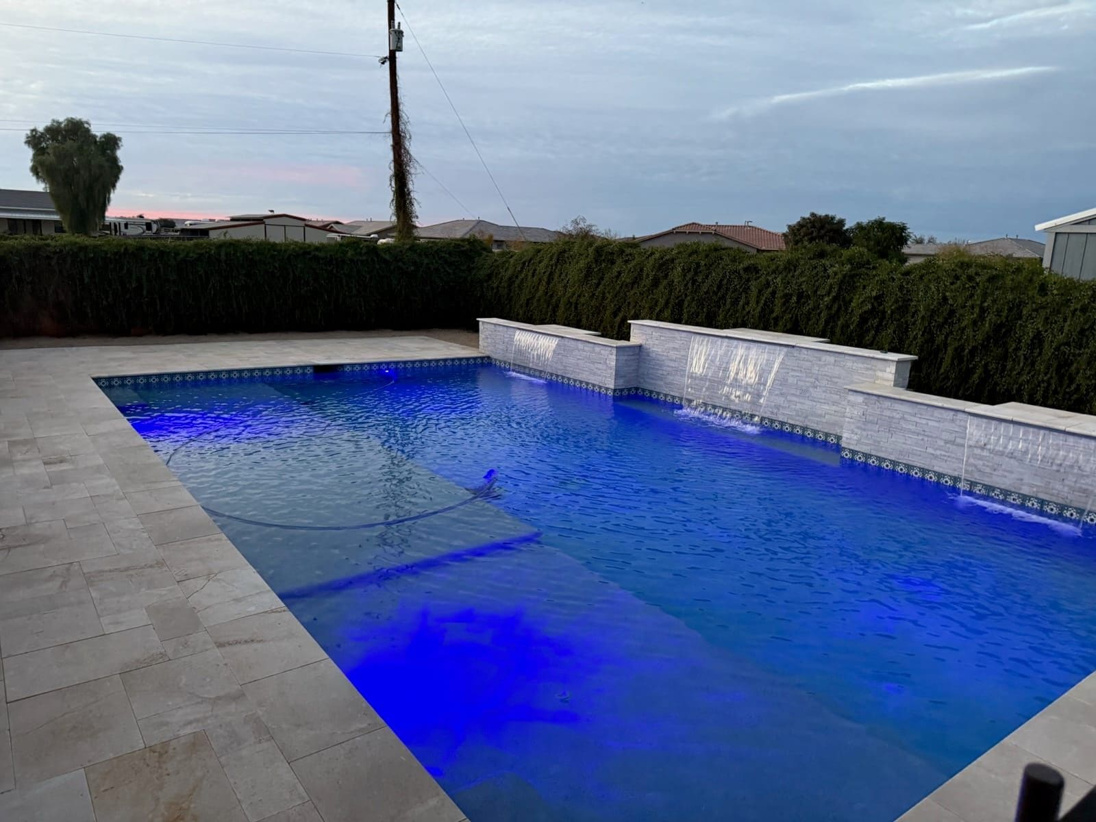 A rectangular pool glowing blue at dusk, with two raised stacked-stone walls pouring sheet waterfalls over patterned waterline tile and a travertine deck.