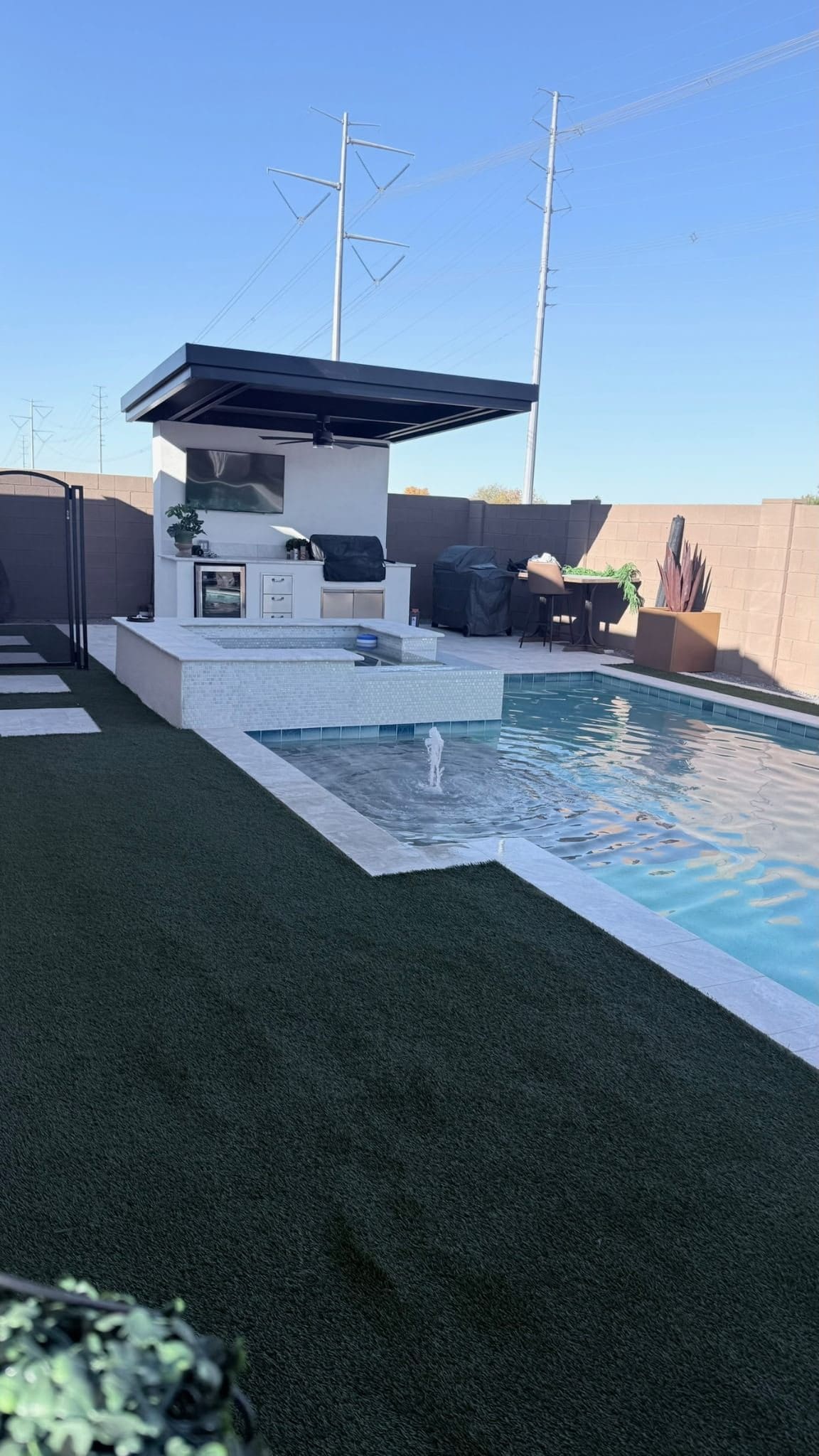 A glass-tile spa and pool with a bubbler fountain beside an outdoor kitchen with a TV under a black cantilevered pergola, framed by artificial turf.