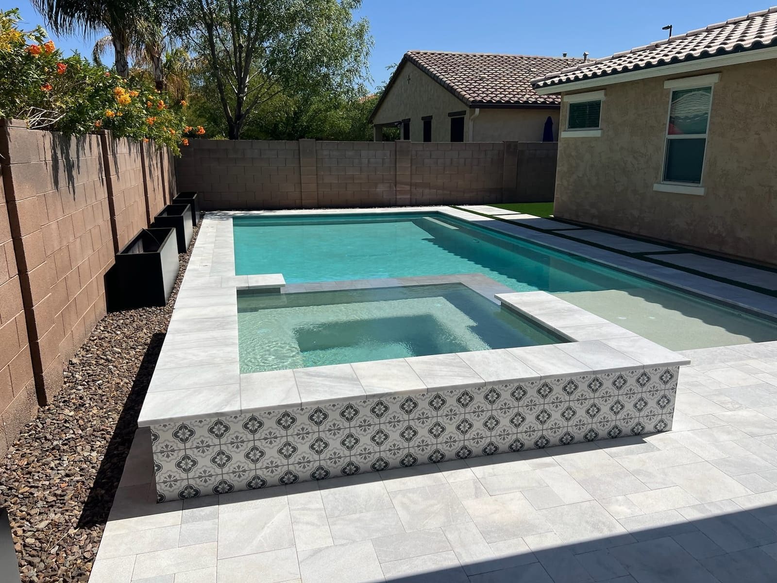 A pool and raised spa faced in patterned cement tile, with light stone coping, gravel planting beds and a block privacy wall.