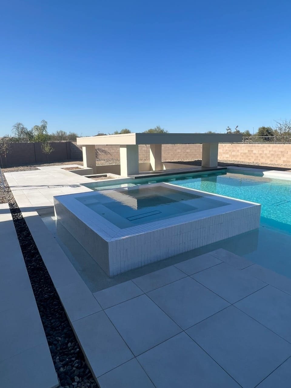 A square raised spa fully clad in white tile at the edge of a bright blue pool, with a shade pavilion and block wall behind.